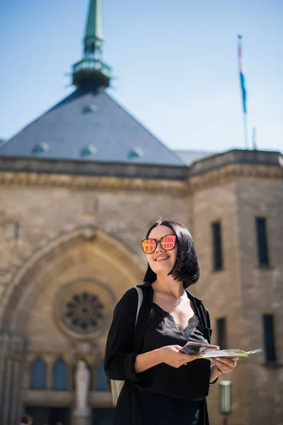 Morena joven turista sosteniendo un mapa de la ciudad y buscando la dirección correcta de pie al aire libre en el entorno urbano.Alegre viajero femenino con gafas de sol haciendo ruta disfrutando de viaje de fin de semana . — Foto de Stock
