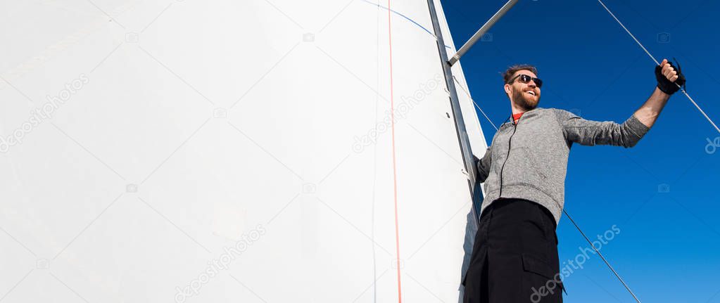 Yacht captain with a beard stands on sail boom on a sailing yacht, holding the rope in his hand and smiling, feeling happy. Adult yachtsman travelling around the world. Copy space