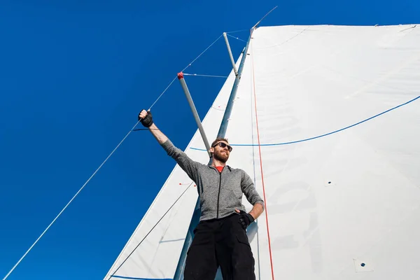 Fiatal férfi áll yacht-boom, és várakozással. Ő tartja a árboc kötél kézzel. A fiatalember jelent. Szürke kapucnis felső-fekete nadrágot visel.. — Stock Fotó