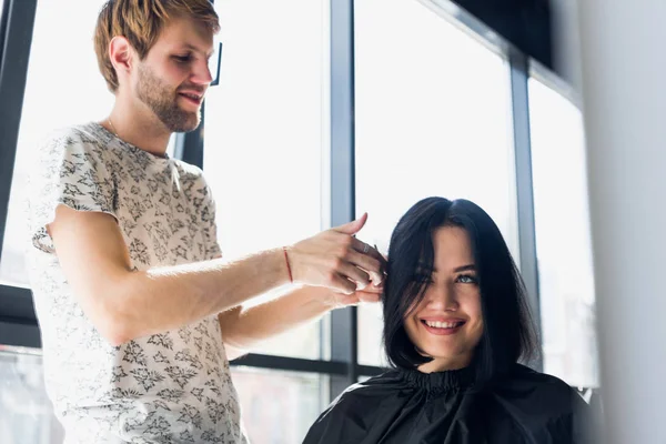 Mulher bonita nova que tem seu cabelo cortado nos cabeleireiros. Jovem cabeleireiro masculino sorrindo e fazendo penteado para o cliente . — Fotografia de Stock