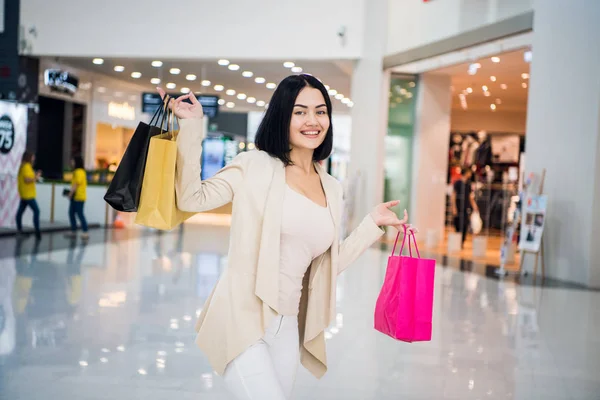 Mooi meisje in winkelcentrum, kijken naar camera, breed glimlachend en kleurrijke boodschappentassen in verhoogde handen houden. Een beetje opzij gedraaid. Het dragen van witte jeans — Stockfoto