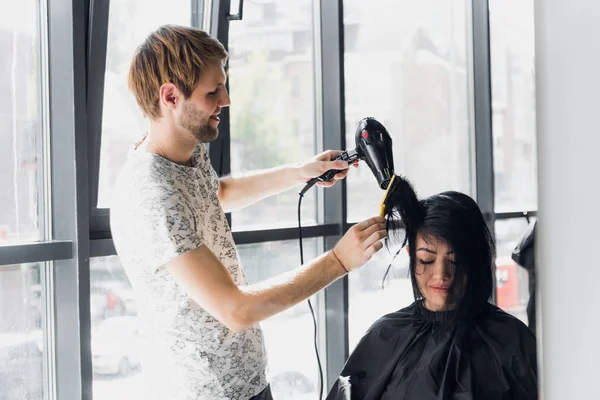 Retrato de cabeleireiro feliz segurando pente e secador de cabelo no salão — Fotografia de Stock