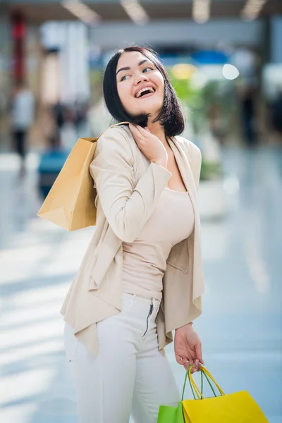 Egy sötét hajú nő visel tompa, gyengéd színekkel rendelkezik, színes, mintás bevásárló táskák sétál egy exkluzív shopping mall. — Stock Fotó