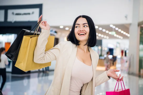 Una mujer de cabello oscuro que usa colores suaves y silenciosos sostiene coloridas bolsas de compras con dibujos en un exclusivo centro comercial . — Foto de Stock