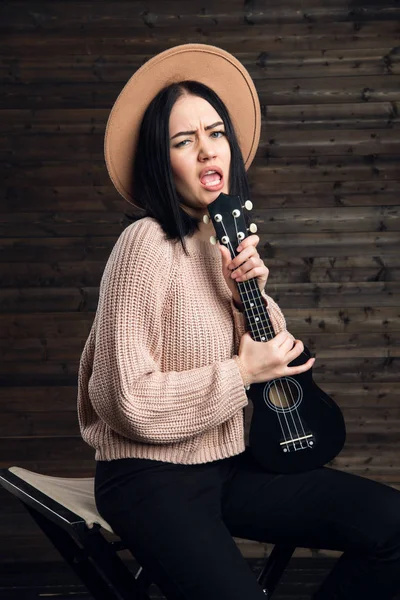 Νέοι αστεία hipster κορίτσι διασκεδάζοντας και παίζοντας την κιθάρα μικρός γιουκαλίλι, τραγουδώντας και χορεύοντας. φοράει vintage καπέλο, χαρά, θετική διάθεση. Φόντο σκοτεινό ξύλινο τείχος. — Φωτογραφία Αρχείου
