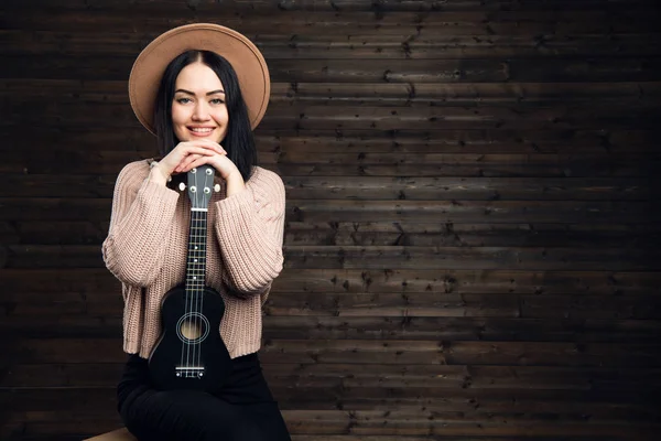 Νεαρή όμορφη γυναίκα που κάθεται σε μια καρέκλα με γιουκαλίλι φορώντας ένα καπέλο. Το κορίτσι με τα χέρια διπλωμένα σε μια μικρή κιθάρα σε σκούρο φόντο ξύλινη. — Φωτογραφία Αρχείου