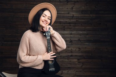 Genç güzel kadın bir şapka giymiş ukulele ile bir sandalyede oturuyor. Koyu ahşap arka plan küçük bir gitar üzerinde katlanmış ellerle kız.