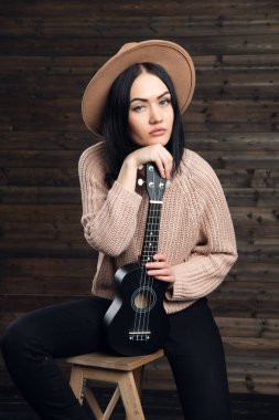 Genç güzel kadın bir şapka giymiş ukulele ile bir sandalyede oturuyor. Koyu ahşap arka plan küçük bir gitar üzerinde katlanmış ellerle kız.