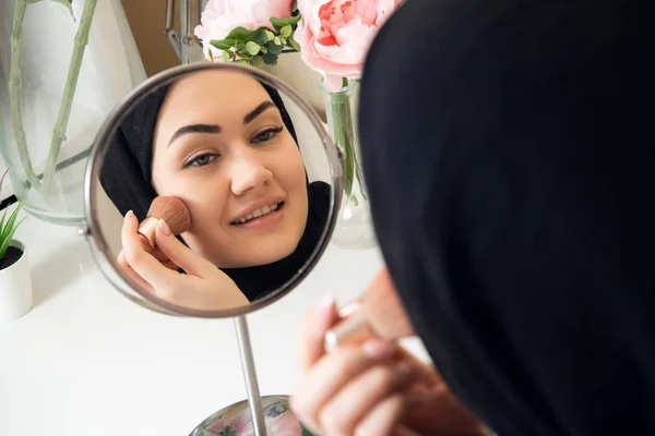 美丽的穆斯林妇女与盖头应用化妆。美丽的女孩看着镜子和应用化妆品。女孩在颧骨上脸红. — 图库照片