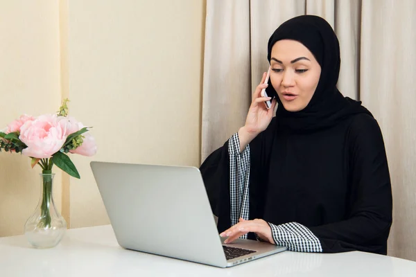 Mujer musulmana hablando por teléfono mientras trabaja con computadora portátil . — Foto de Stock