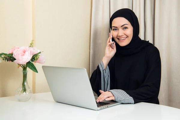 Mujer musulmana hablando por teléfono mientras trabaja con computadora portátil . — Foto de Stock