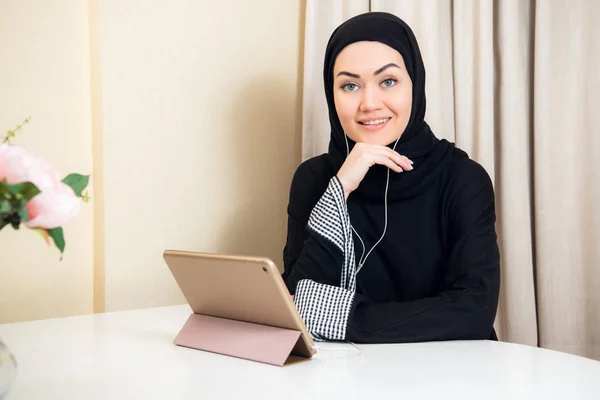 Mujer musulmana sentada en casa con tableta, con auriculares. Descanse en casa, preparándose para ver una película o escuchar una música . — Foto de Stock
