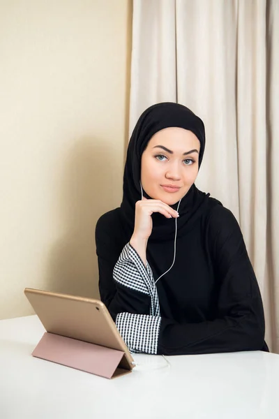 Mujer musulmana sentada en casa con tableta, con auriculares. Descanse en casa, preparándose para ver una película o escuchar una música . — Foto de Stock