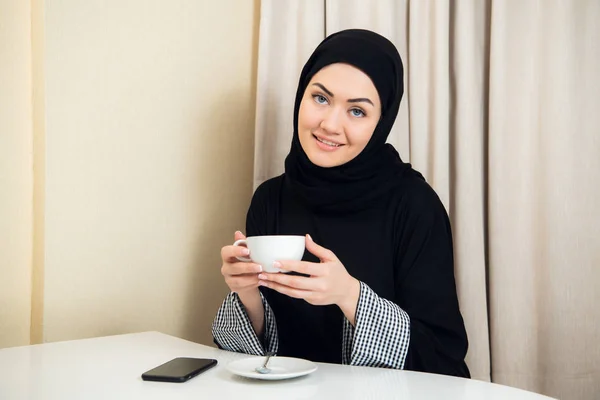 portrait of attractive asian woman wearing hijab enjoying coffee and smiling to camera