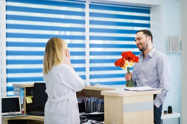 Hermosa enfermera coqueteando con un apuesto cliente masculino. Le trajo flores al trabajo. . — Foto de Stock