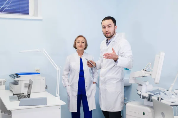 Médico sênior conversando com o jovem assistente de pé no escritório ginecológico com cadeira e lâmpada no fundo — Fotografia de Stock