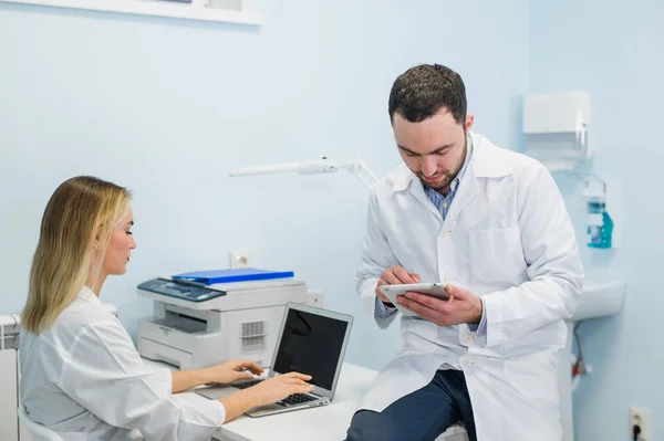 Due colleghi medici sono seduti sul posto di lavoro e utilizzano la tecnologia moderna per il loro lavoro, in camici bianchi, concentrati — Foto Stock