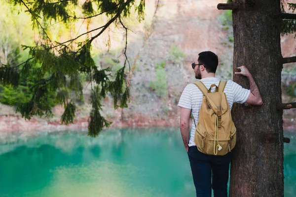 Stilig man över grön natur och blå sjön står nära trädet. Äventyr, Resor, turism och fritid koncept - ung pojke avkopplande i skogen. — Stockfoto