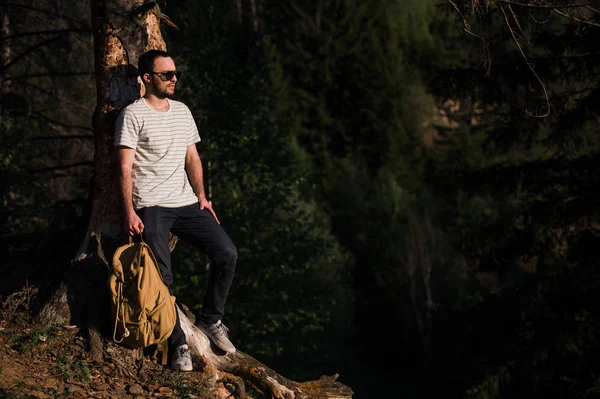 Jonge knappe aantrekkelijke bebaarde model man met rugzak in het bos in de buurt van boom poseren. — Stockfoto