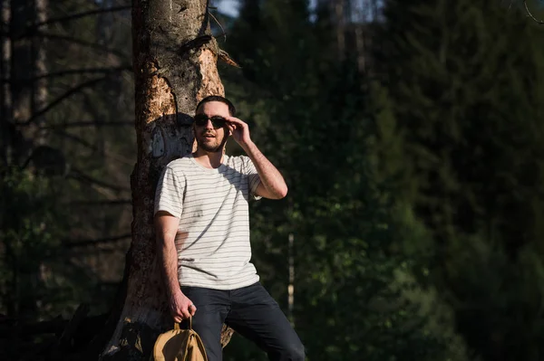 Young handsome attractive bearded model man with backpack in the woods posing near tree.