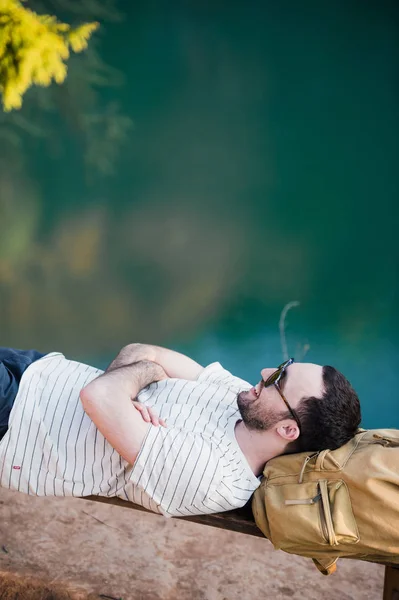 Человек отдыхает во время похода, лежа на деревянной скамейке в лесу. Пешие прогулки на природе — стоковое фото