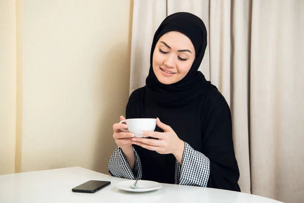 Arab women in hijab holding and drinking coffee cup sitting at home