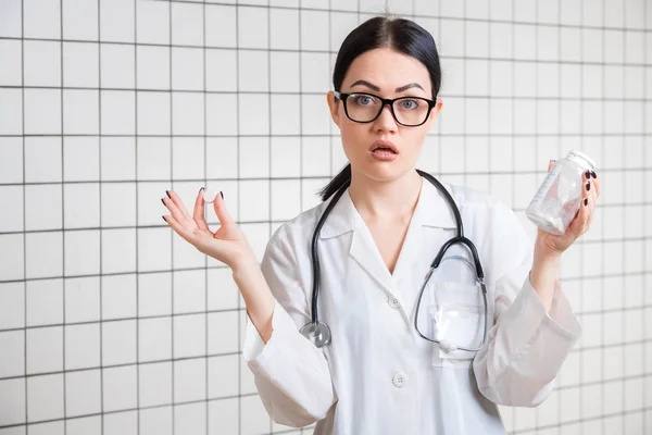 Sorpresa medico femminile gesticolando con le mani e le pillole mentre in piedi con la bocca aperta su sfondo studio medico . — Foto Stock