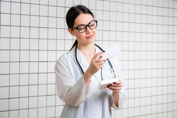 Una simpatica ragazza carina con capelli scuri e occhiali, indossando un camice da laboratorio, legge attentamente le informazioni sulla confezione in una farmacia moderna.. — Foto Stock