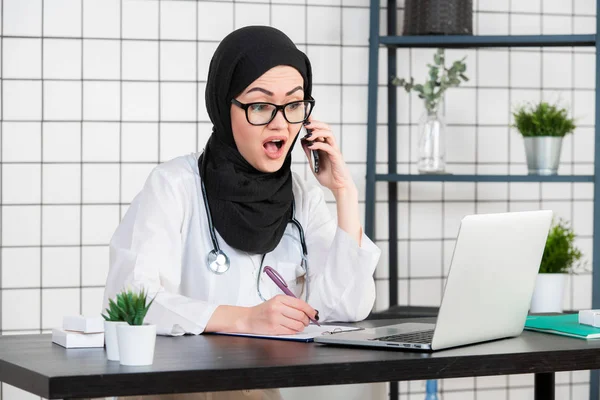 책상에 앉아 랩탑을 보고 있는 여성 베일을 쓴 과학자가 휴대폰으로 통화하면서 충격적 인 얼굴 감정으로 입을 연다.. — 스톡 사진