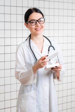 Genç, zayıf, koyu renk saçlı, gözlüklü bir kız, tıbbi bir kıyafet giymiş, eczanenin arka planındaki ilaç paketinin yanında bir şeyler okuyor..