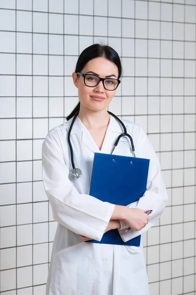 Young beautiful female doctor in white surgical coat with black stethoscope and blue paper holder in hands standing at medical office