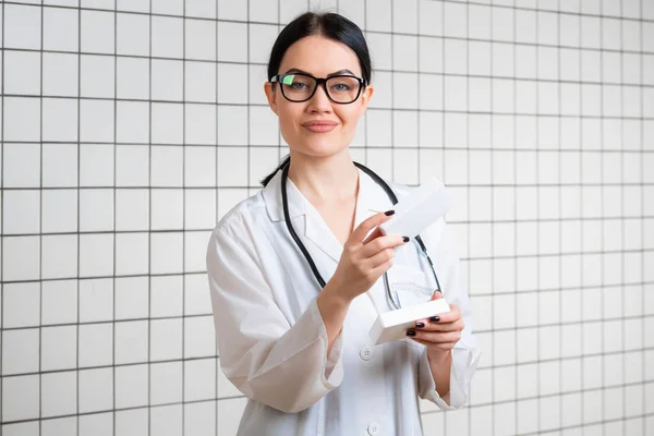 Una giovane ragazza dai capelli scuri con gli occhiali, vestita in generale medica, legge qualcosa sul pacchetto di pillole accanto allo sfondo dell'ufficio in farmacia.. — Foto Stock