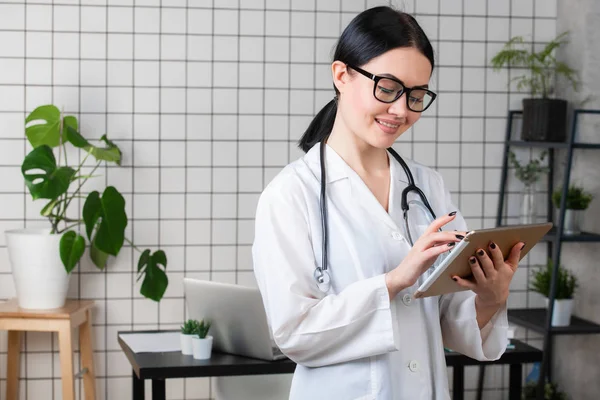 Médico morena femenina usa gafas usando tableta, mujer inteligente y encantadora con el gadget y estetoscopio en el fondo blanco de la oficina — Foto de Stock
