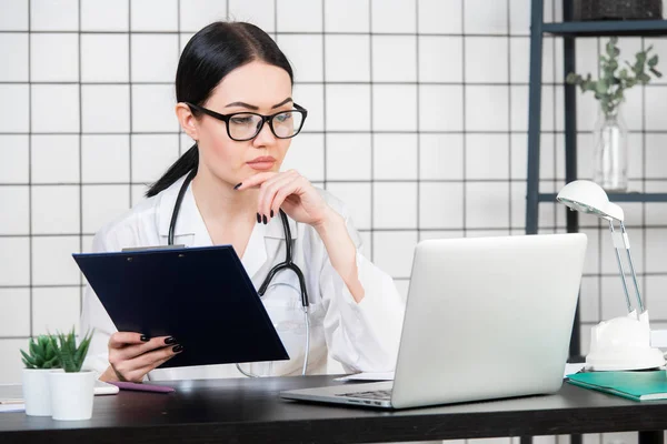 Médico morena femenina usa gafas usando tableta, mujer inteligente y encantadora con el gadget y estetoscopio en el fondo blanco de la oficina — Foto de Stock