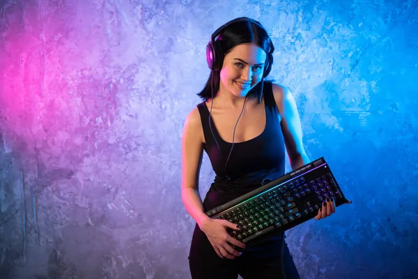 Νέοι σέξι ντίσκο κορίτσι φορώντας ακουστικά τυχερού παιχνιδιού κρατώντας Gaming Kyeboard. Δωμάτιο αναμμένα στο ρετρό, Retrowave ή Cyberpunk στυλ με νέον και ροζ φώτα. — Φωτογραφία Αρχείου