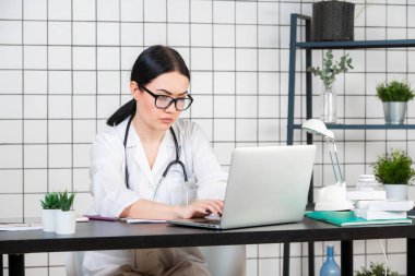 Masasında oturan gülümseyen genç bir kadın hastanedeki hafif büroda tıbbi belgelerle bilgisayarla çalışıyor. Tıbbi önlüklü kadın doktor, danışma odasında steteskop. Sağlık sigortası konsepti.