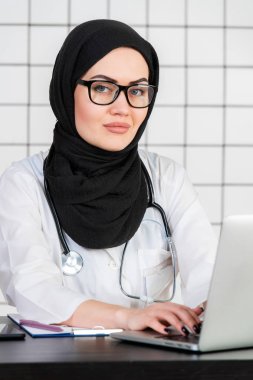 Gözlüklü, steteskoplu ve beyaz bir ofis geçmişine ait belgeleri olan gülümseyen Müslüman bir doktor.