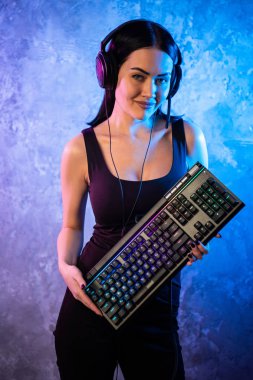 Fotoğraf makinesi içine gaming klavye ve kulaklık ve görünüyor güzel genç Pro Gamer kız ayakta portresi. Çekici Geek Kız oyuncu tarafından Neon ışıkları yaktı odasında gözlüklü.