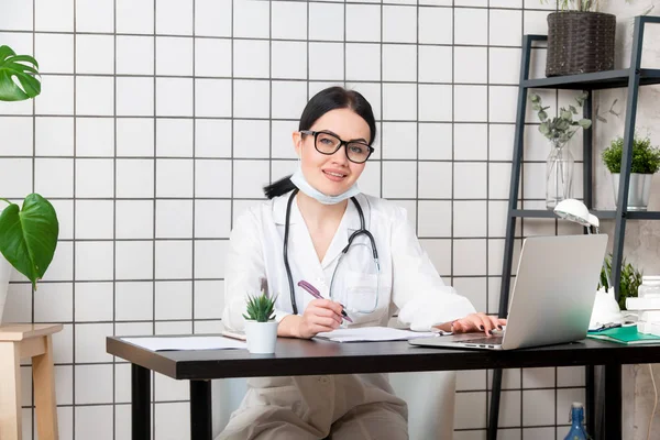 젊은 여성 이 책상에 앉아서 웃으면서 병원에 있는 밝은 사무실에서 의학 문서들을 가지고 컴퓨터를 연구하고 있습니다. 의료 복을 입은 여자 의사, 컨설팅 룸의 청진기. 의료 상의 개념. — 스톡 사진