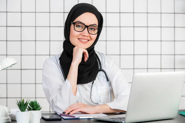 A Muslim Doctor smiling with glasses and stethoscope and documents on a white office background