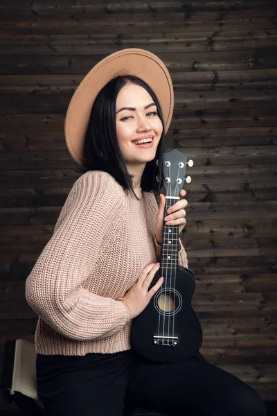 Attraktiv känslomässiga unga kaukasiska kvinna i tröja och hatt spela ukulele. Vacker kvinna sångare spela musikinstrument inomhus. Människor, musik, glädje och nöje — Stockfoto