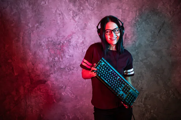 Junges sexy Disco-Mädchen mit Gaming-Headset und Gaming-Tastatur. Raum beleuchtet im Retro-, Retrowellen- oder Cyberpunk-Stil mit Neon- und rosafarbenen Lichtern. — Stockfoto