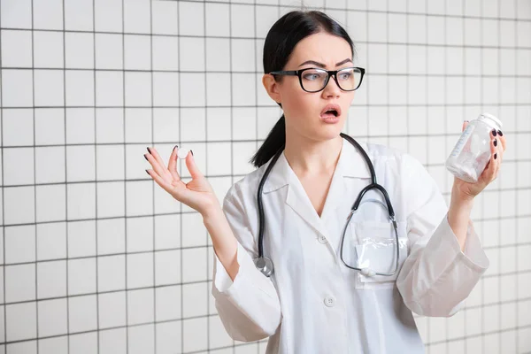 Sorpresa medico femminile gesticolando con le mani e le pillole mentre in piedi con la bocca aperta su sfondo studio medico . — Foto Stock