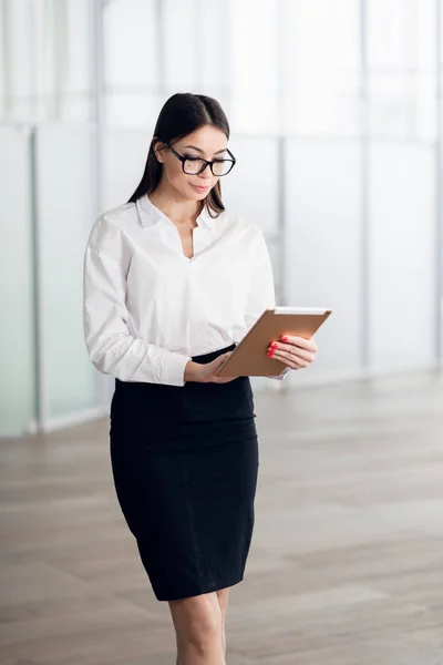 Atractiva joven mujer con gafas y leyendo su tableta de pantalla táctil mientras está de pie dentro del edificio comercial —  Fotos de Stock