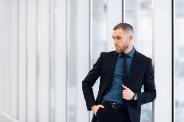 Modern bir takım elbise giyen şık genç bir işadamı, bir ofis binasının en üst katında durmuş, büyük pencerelerden manzaraya bakıyor.