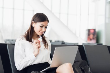 Young business woman with laptop and healthy snack in her hand sitting at departures terminal gate. Traveler waits for his transport connection. Waiting at the airport.