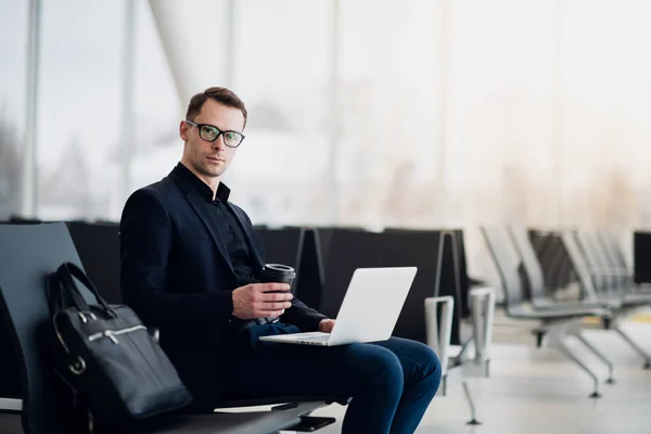 En affärsman sitter vid flygplatsen parken arbetar med sin laptop och dricka takeaway kaffe i väntan på flygningen avgång. — Stockfoto