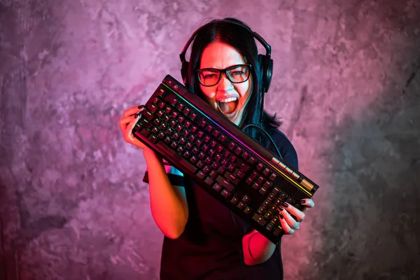 Επαγγελματική Gamer Girl στο MMORPG παιχνίδι στρατηγικής βίντεο. Shes Αυτή θέτει πάνω από πολύχρωμο μπλε και ροζ φόντο με ένα πληκτρολόγιο τυχερών παιχνιδιών. Φοράει ακουστικά παιχνιδιών.. — Φωτογραφία Αρχείου