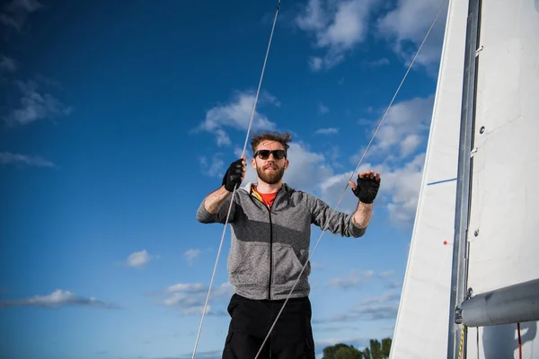 Napszemüveg fiatal tengerész rendelkezik, és mindkét kezével fő vitorla mozog. Ő is boldog és mosolyog. A fiatalember vitorlás yacht készül. A napos, kívül. — Stock Fotó