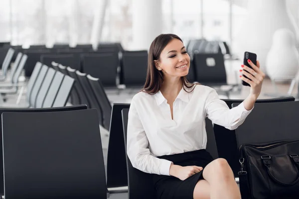 Joven mujer de negocios en el aeropuerto internacional, haciendo selfie con teléfono móvil y esperando su vuelo. Pasajera en la terminal de salida, en interiores. — Foto de Stock